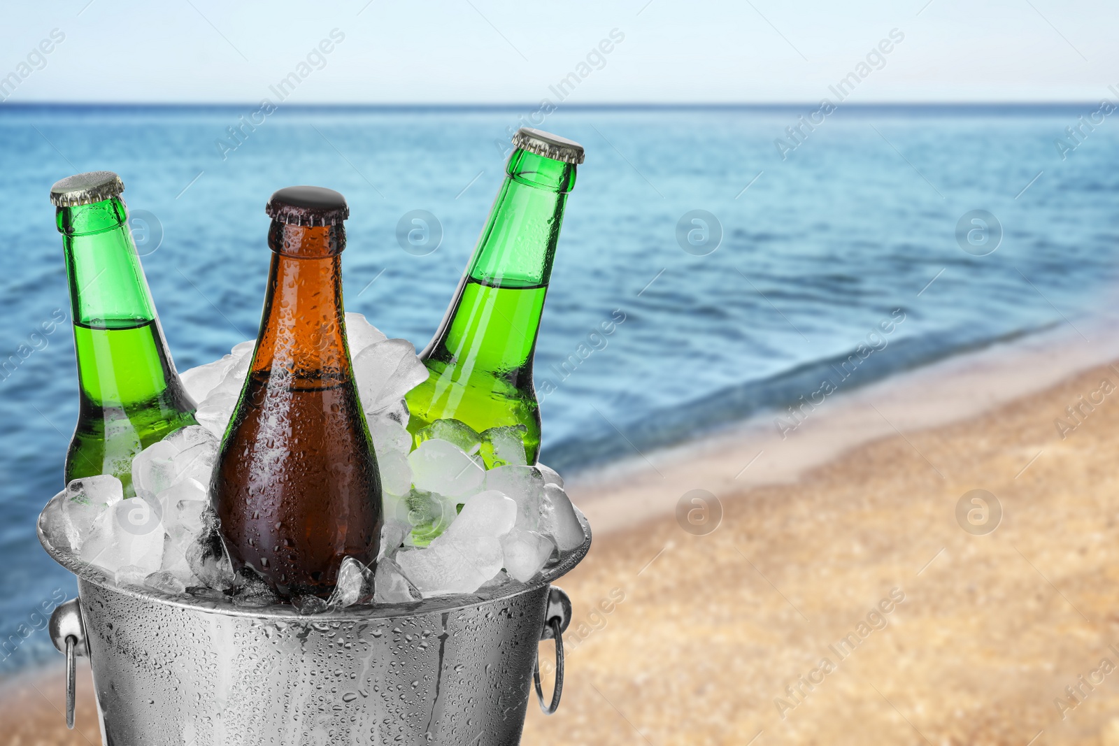 Image of Bottles of beer with ice cubes in metal bucket against blurred sea and sandy beach. Space for text