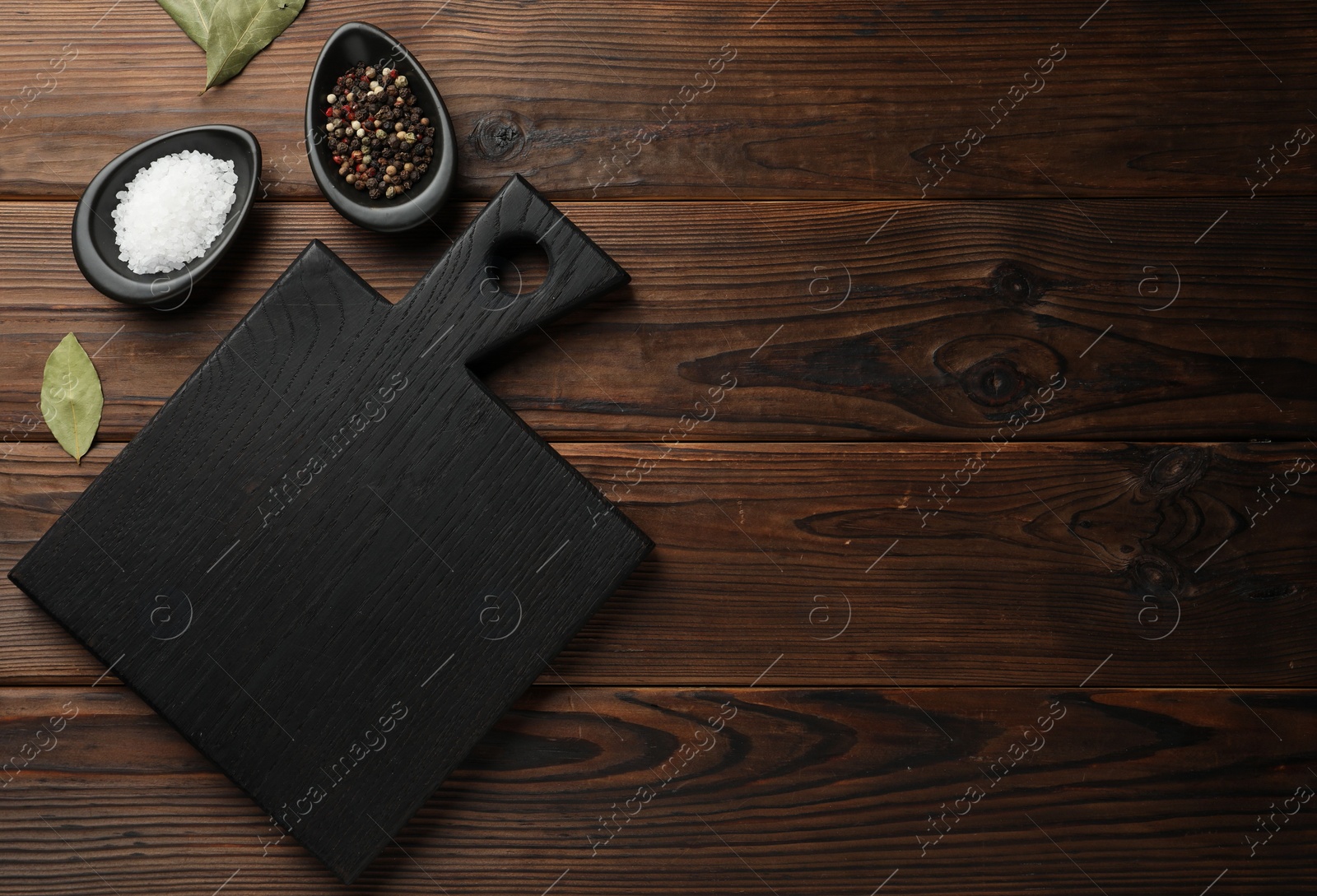 Photo of Black cutting board, salt, pepper and bay leaves on wooden table, flat lay. Space for text