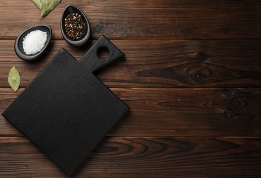 Photo of Black cutting board, salt, pepper and bay leaves on wooden table, flat lay. Space for text