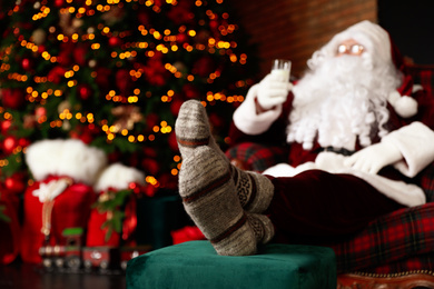 Photo of Santa Claus with glass of milk resting in armchair near Christmas tree, focus on legs