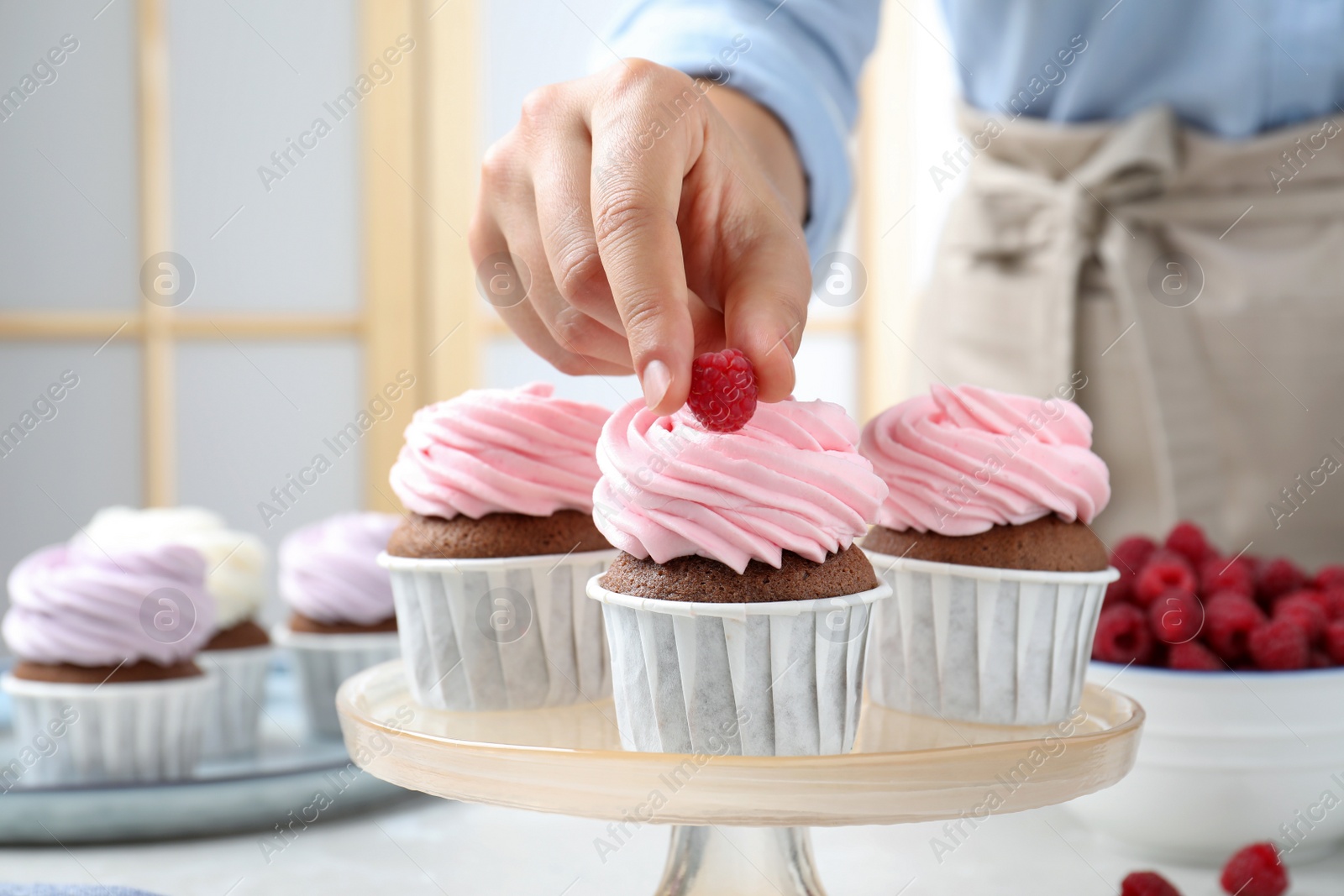 Photo of Woman decorating delicious cupcakes with fresh raspberries at table indoors, closeup