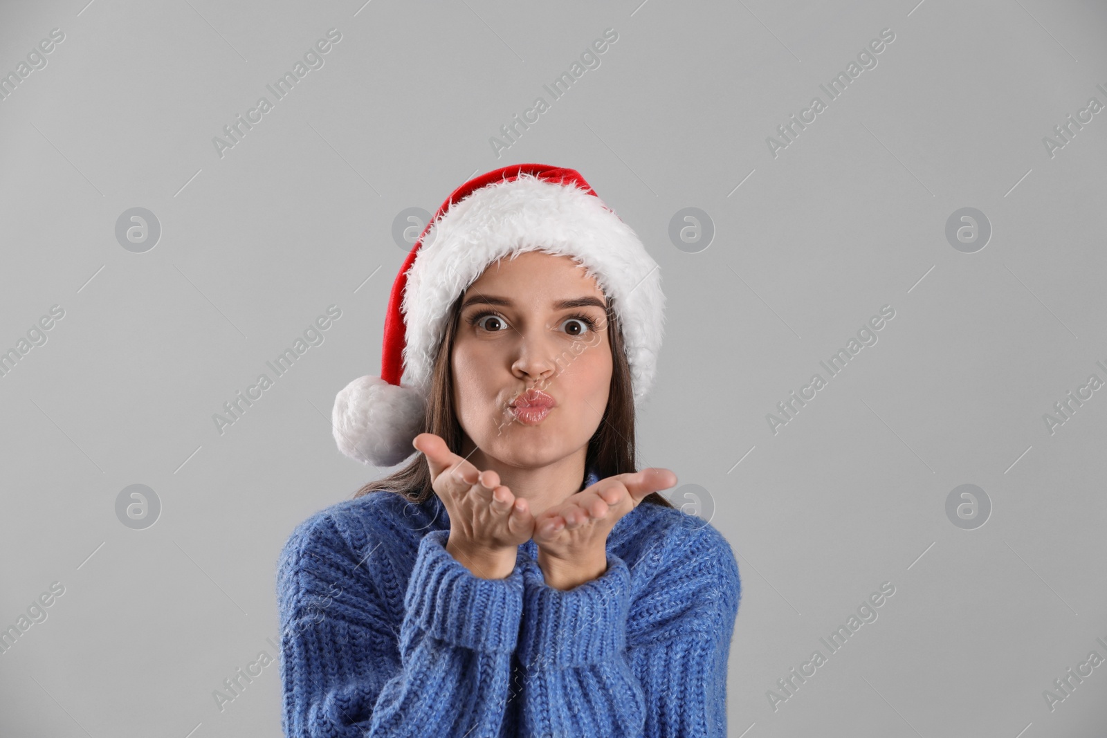 Photo of Pretty woman in Santa hat and blue sweater blowing kiss on grey background