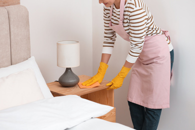 Photo of Young chambermaid wiping dust from nightstand in bedroom, closeup