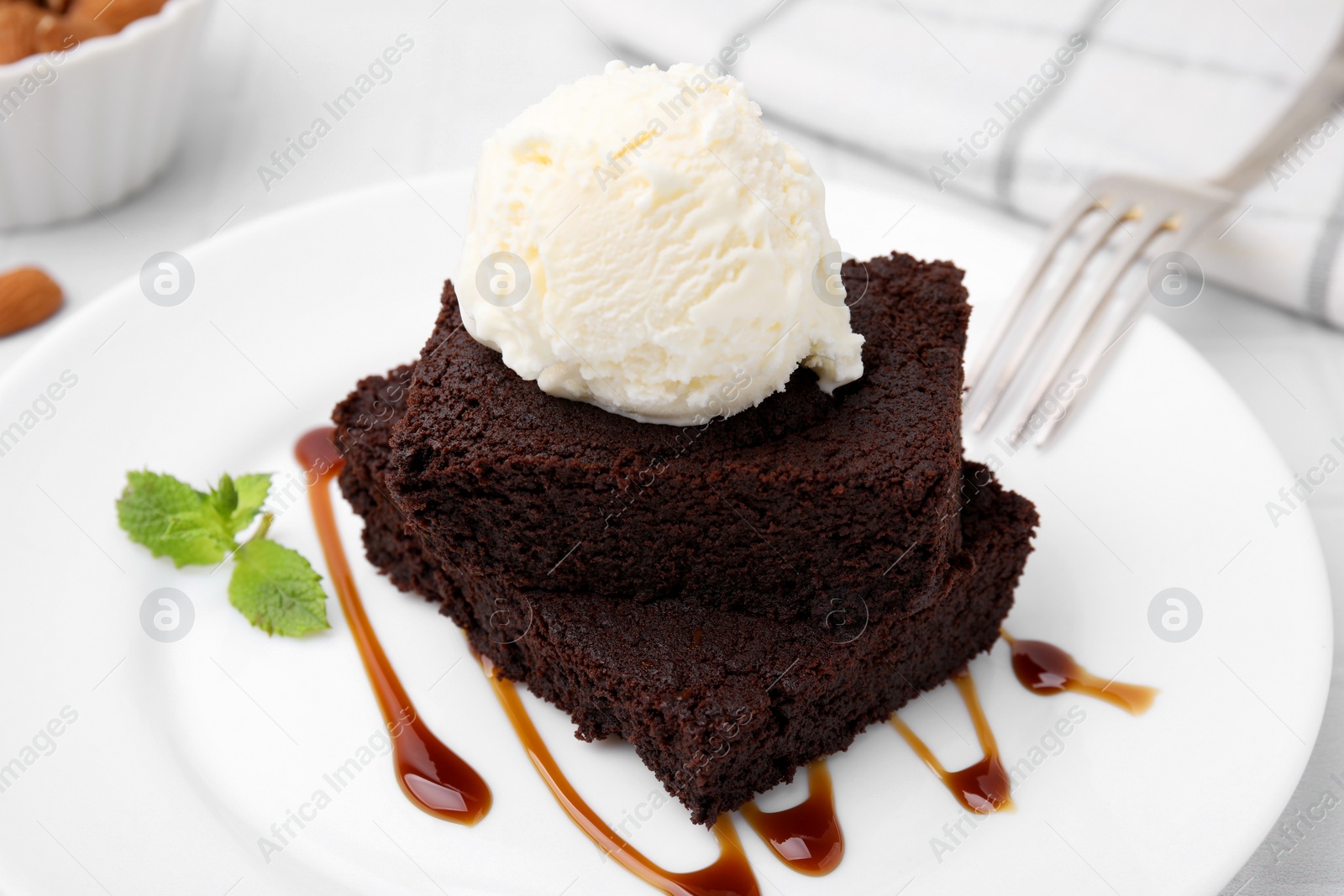 Photo of Tasty brownies served with ice cream and caramel sauce on white table, closeup