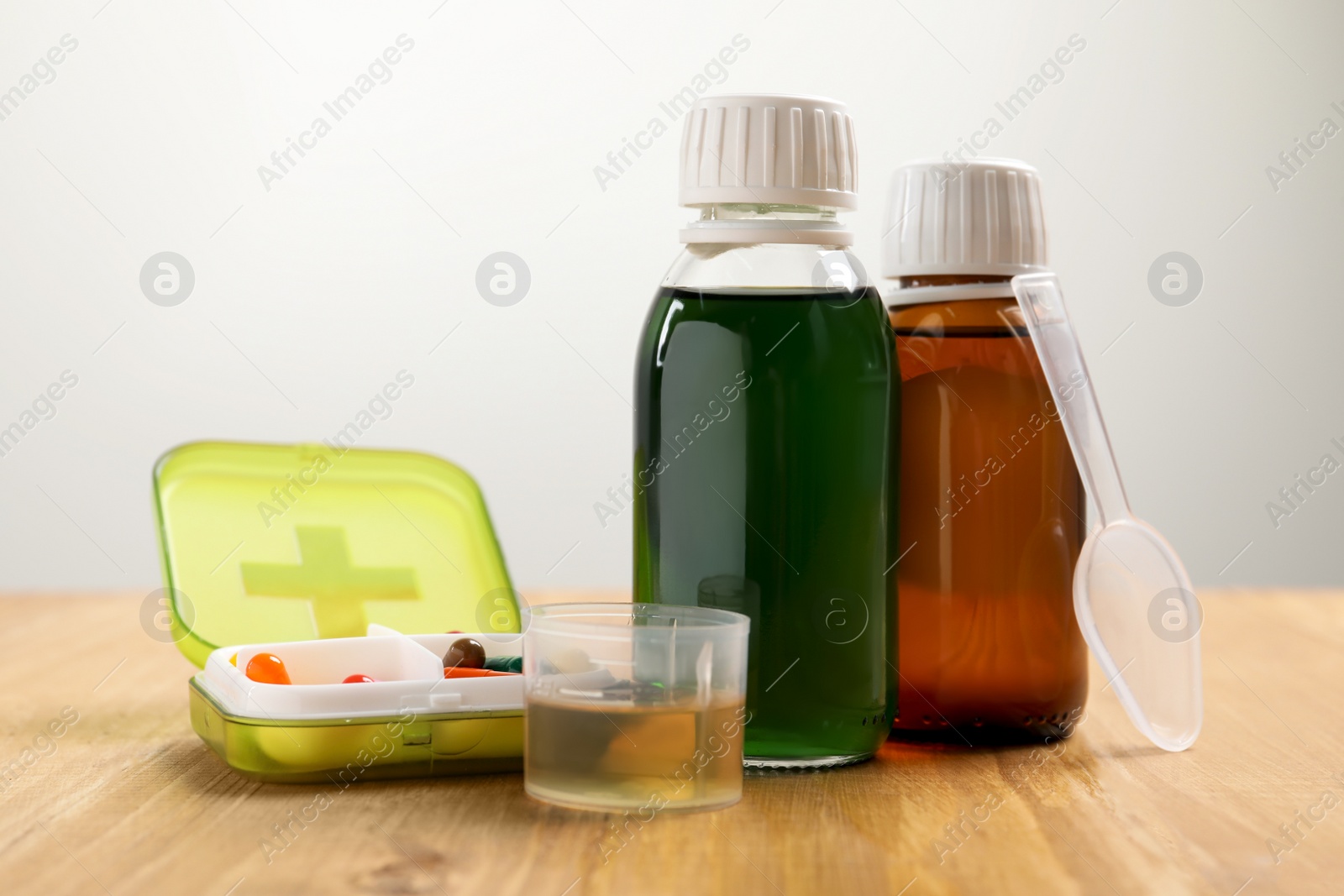 Photo of Bottle of syrup, measuring cup, dosing spoon and pills on wooden table against light background. Cold medicine