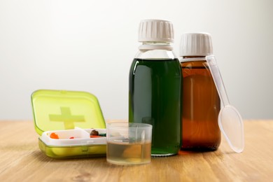 Bottle of syrup, measuring cup, dosing spoon and pills on wooden table against light background. Cold medicine