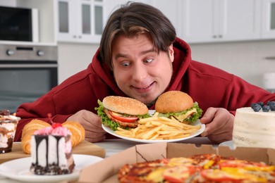 Photo of Hungry overweight man holding plate with tasty burgers and French fries at table in kitchen
