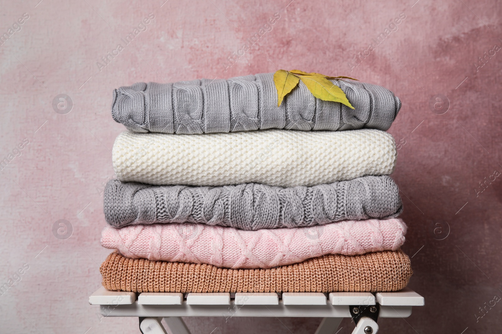 Photo of Stack of warm knitted clothes and autumn leaves on white wooden table against pink background