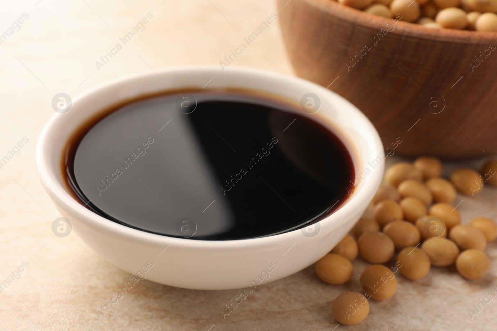 Photo of Soy sauce in bowl and soybeans on beige table, closeup