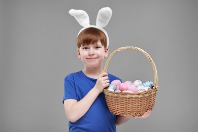 Easter celebration. Cute little boy with bunny ears and wicker basket full of painted eggs on grey background