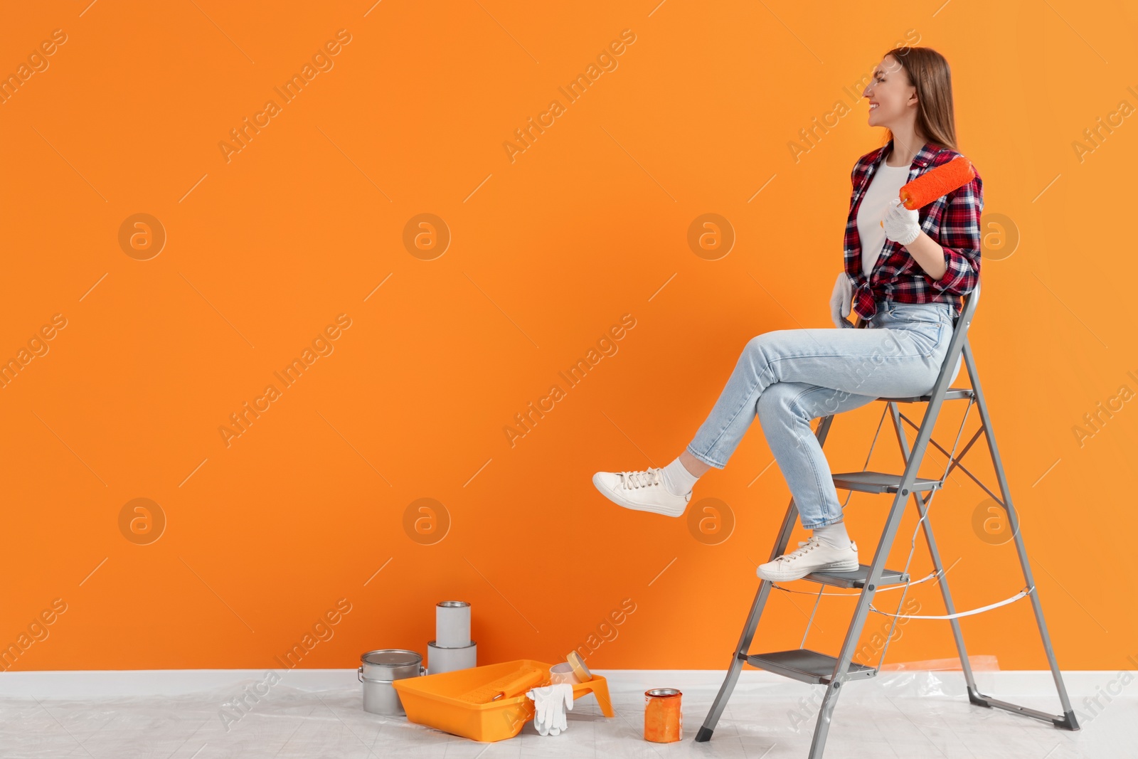 Photo of Designer with roller sitting on folding ladder near freshly painted orange wall indoors, space for text