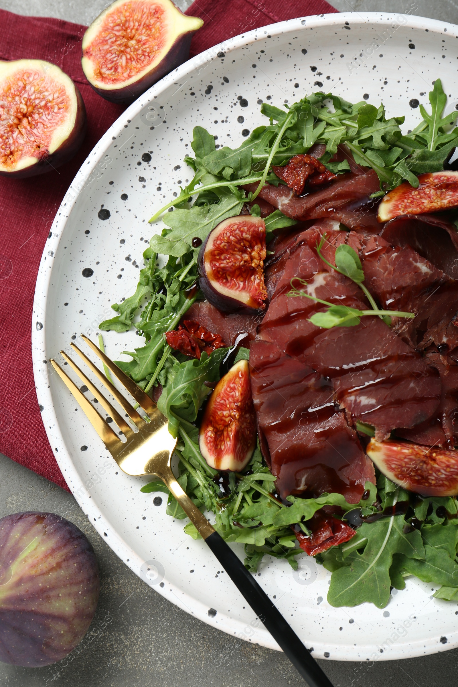Photo of Plate of tasty bresaola salad with figs, sun-dried tomatoes, balsamic vinegar and fork on grey table, flat lay