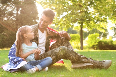 Father in military uniform with American flag and his little daughter sitting on grass at park