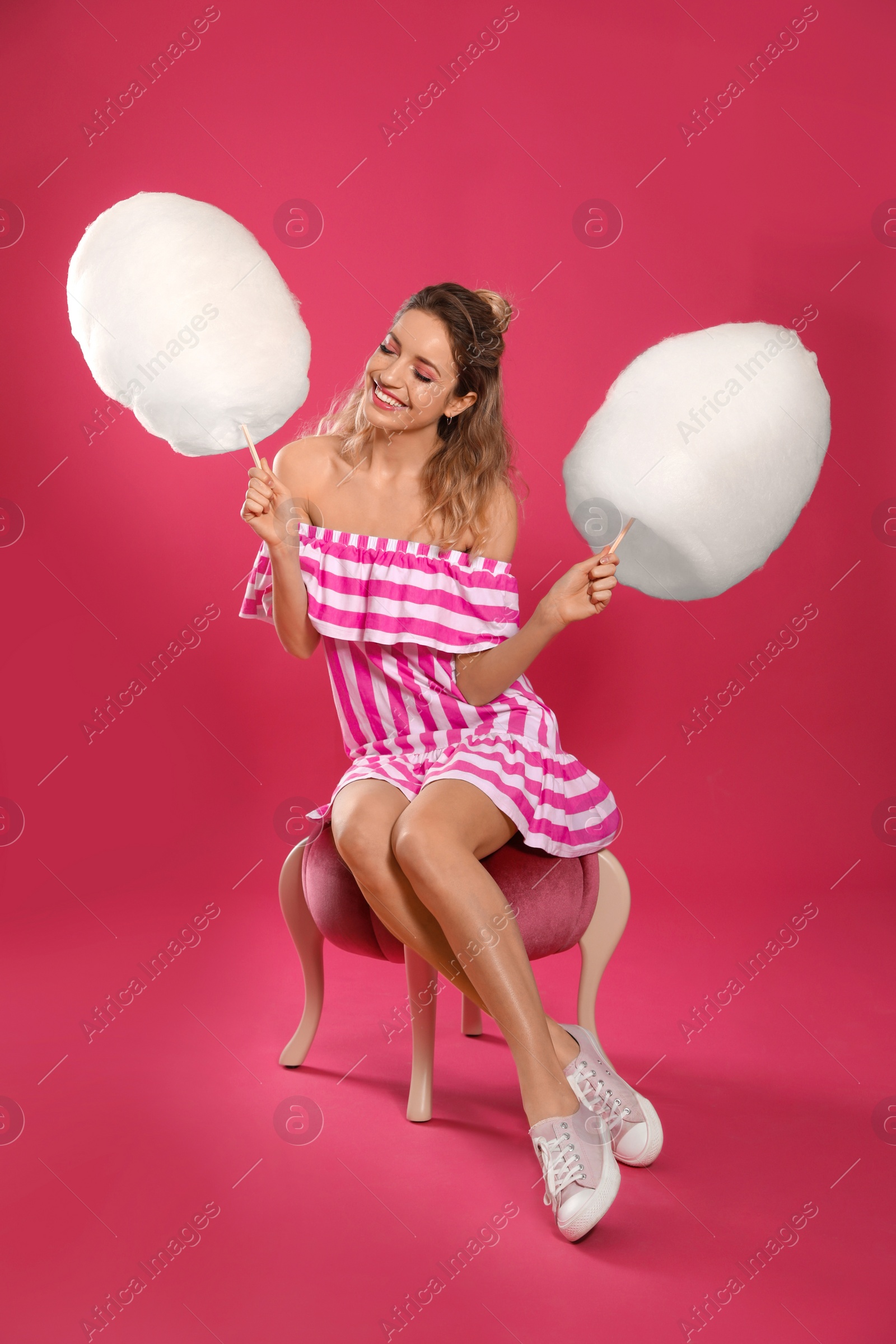 Photo of Full length portrait of young woman with tasty cotton candy sitting on pouf, pink background
