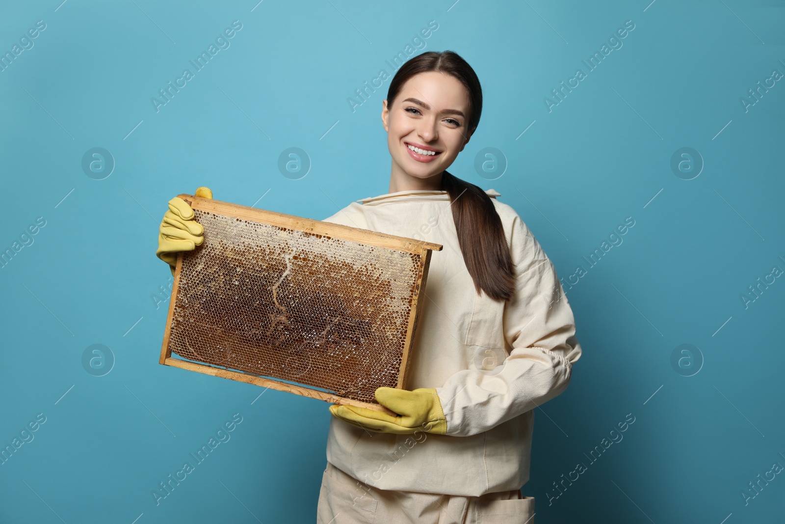 Photo of Beekeeper in uniform holding hive frame with honeycomb on light blue background