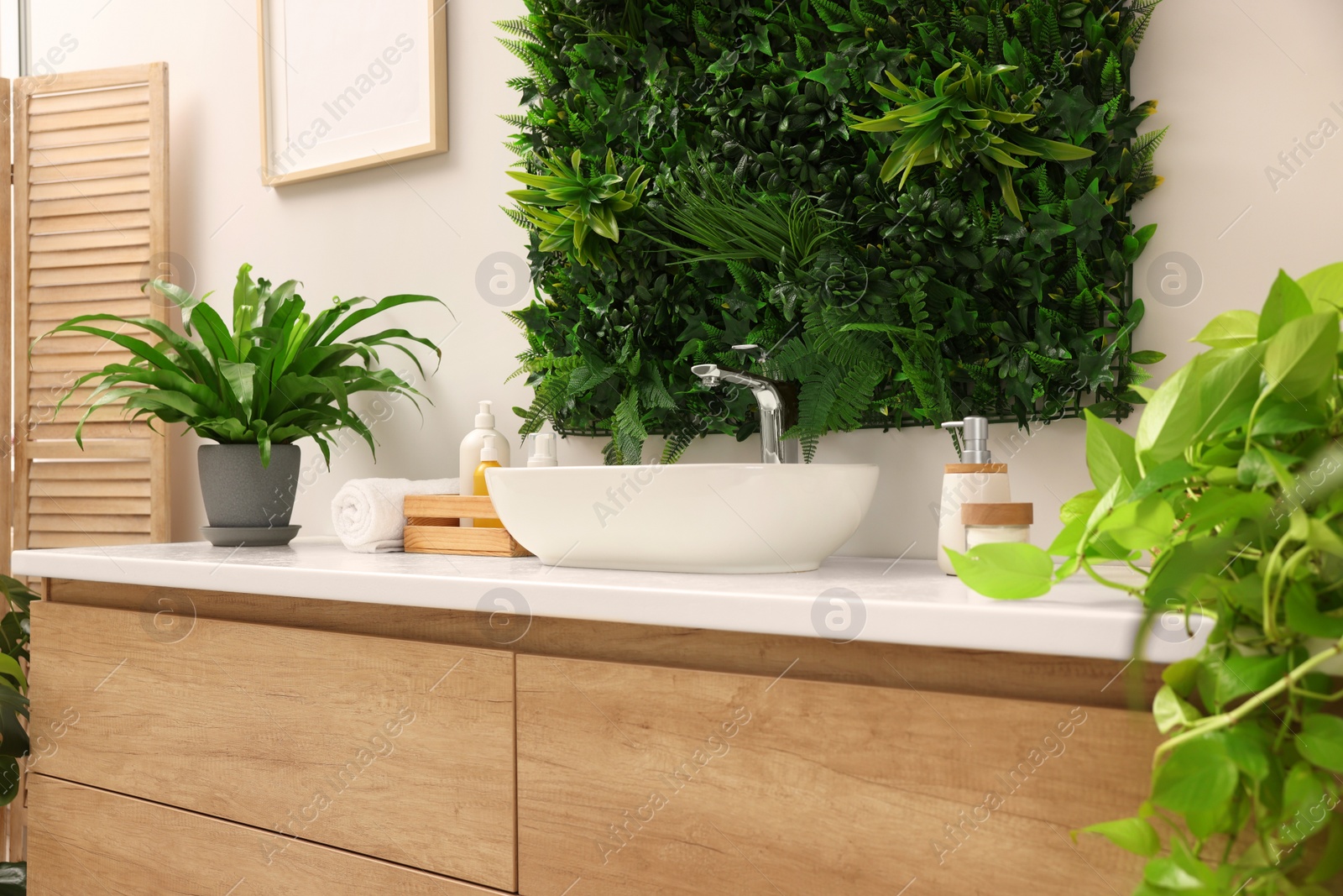 Photo of Green artificial plants, vessel sink and different personal care products in bathroom