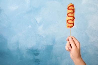 Woman holding delicious corn dog with ketchup on light blue background, closeup. Space for text