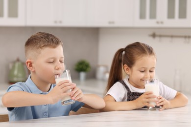 Cute children drinking fresh milk from glasses at white table in kitchen