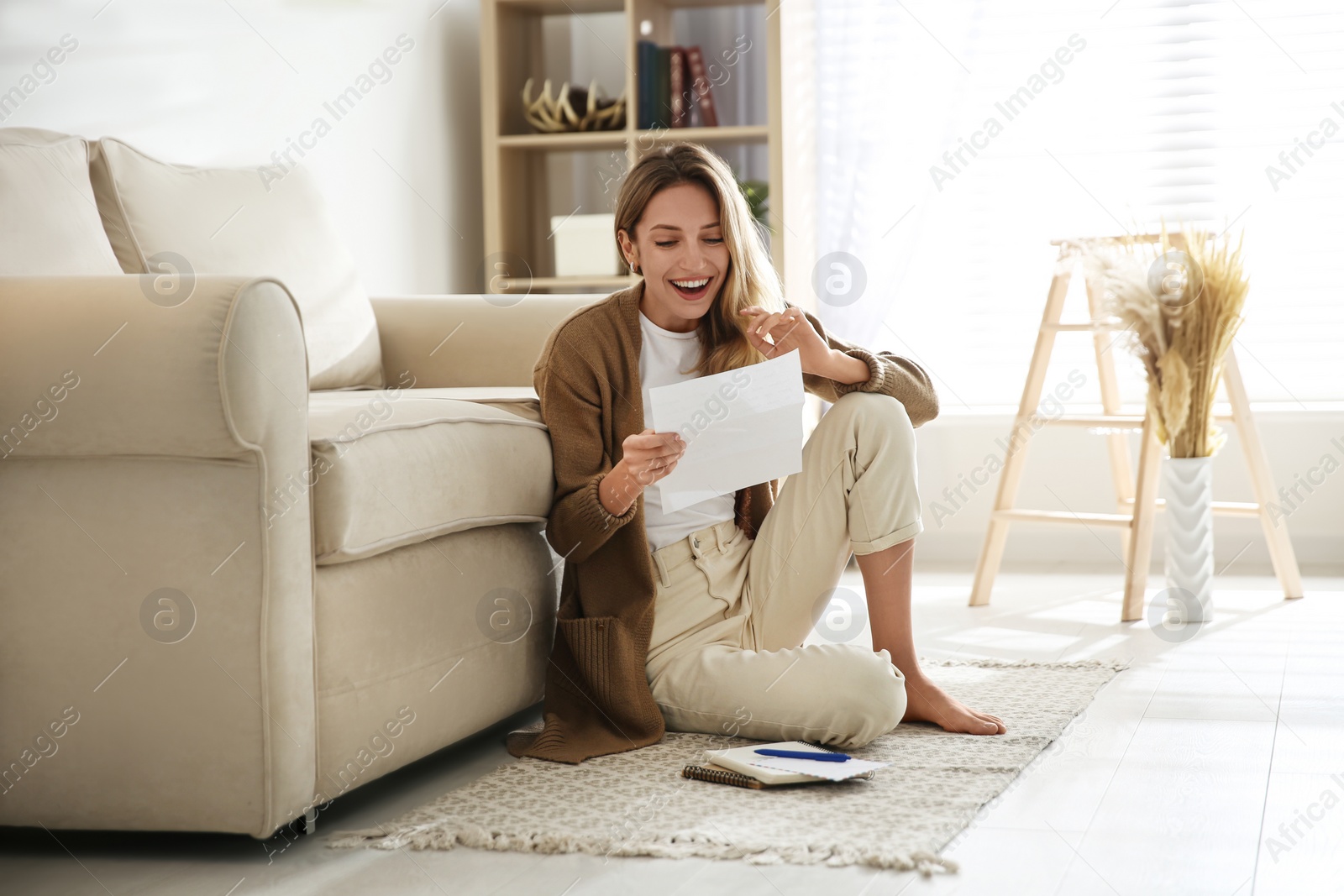 Photo of Happy woman reading letter while sitting on floor near sofa at home