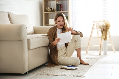 Photo of Happy woman reading letter while sitting on floor near sofa at home