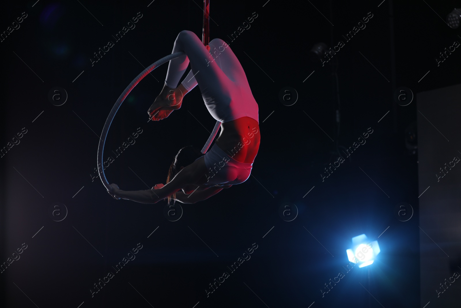 Photo of Young woman performing acrobatic element on aerial ring against dark background