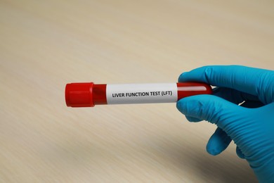 Laboratory worker holding tube with blood sample and label Liver Function Test at beige table, closeup