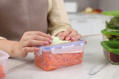 Photo of Woman sticking paper note onto container with fresh carrots at white marble table in kitchen, closeup. Food storage