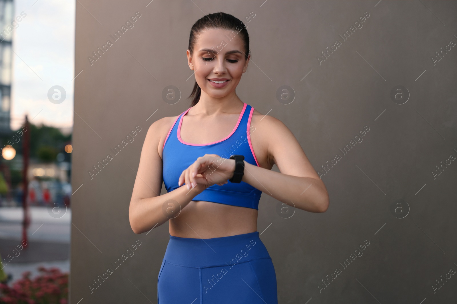 Photo of Beautiful woman in stylish sportswear checking smart watch near black wall outdoors