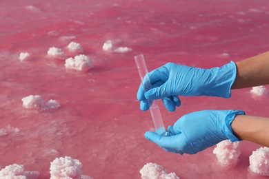 Photo of Laboratory worker with test tube taking sample from pink lake for analysis, closeup