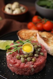 Photo of Tasty beef steak tartare served with quail egg and other accompaniments on black plate, closeup