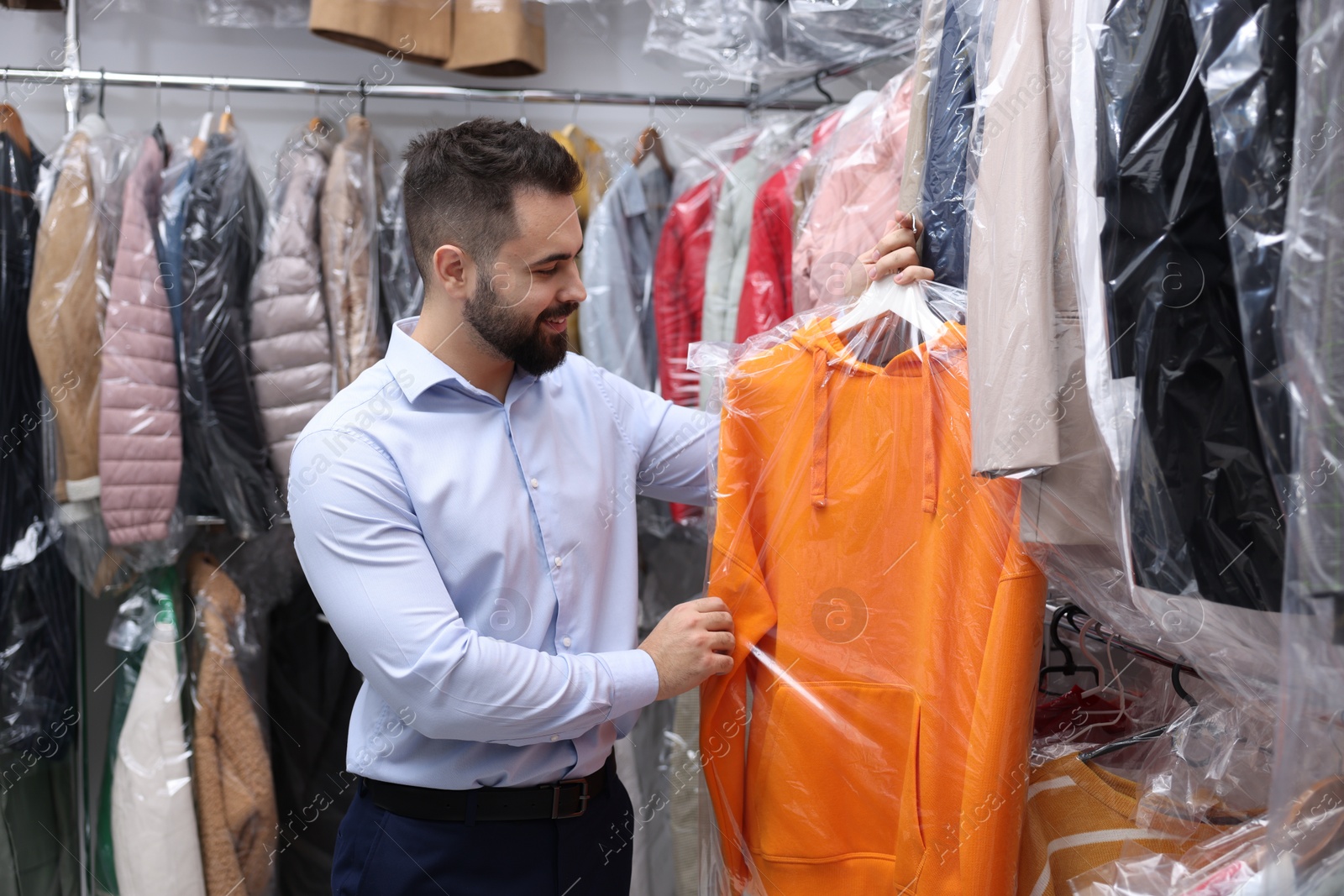 Photo of Dry-cleaning service. Happy worker holding hanger with hoodie in plastic bag near other clothes indoors