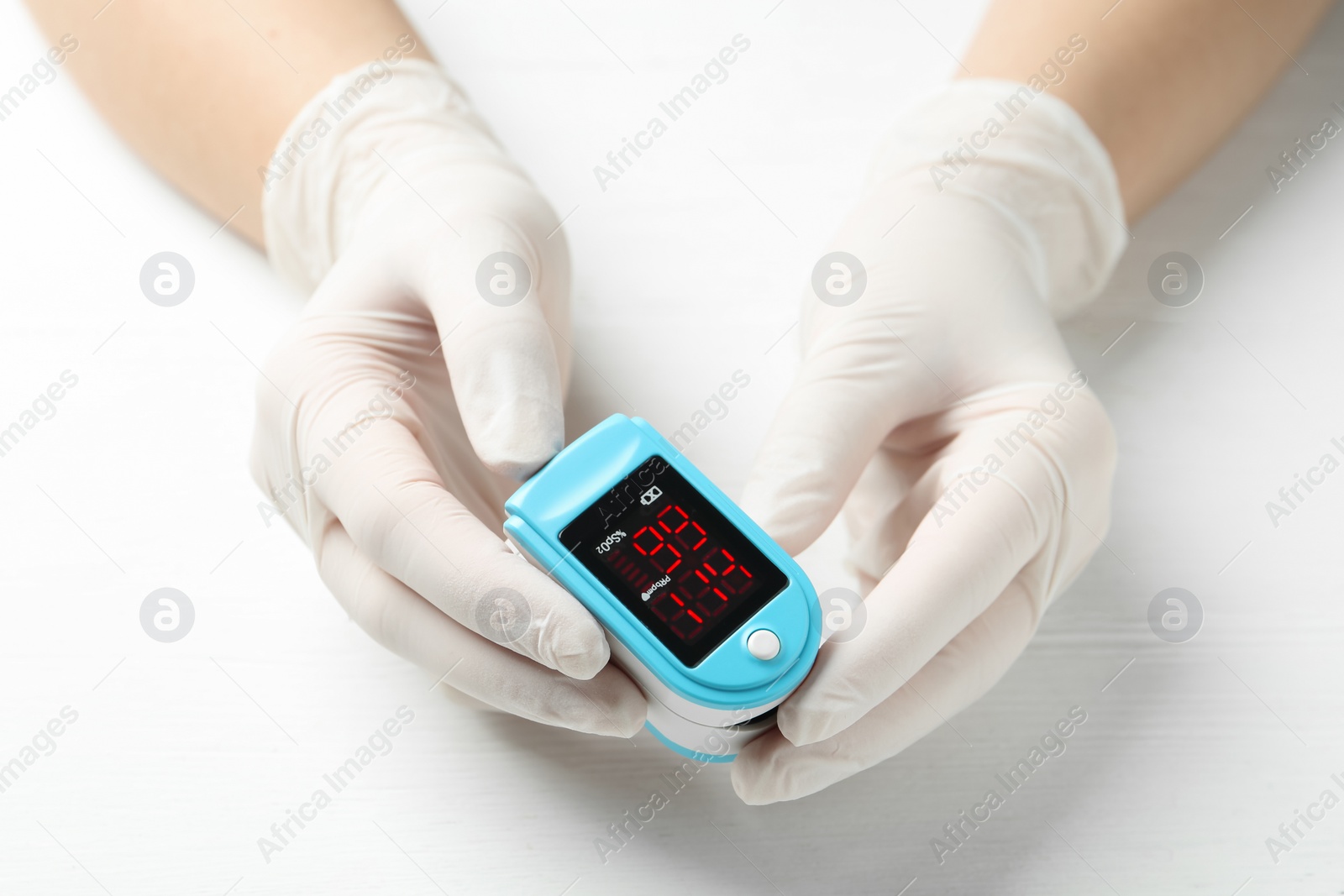 Photo of Doctor in gloves holding fingertip pulse oximeter at white wooden table, closeup
