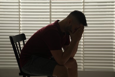 Sad man sitting near closed blinds indoors