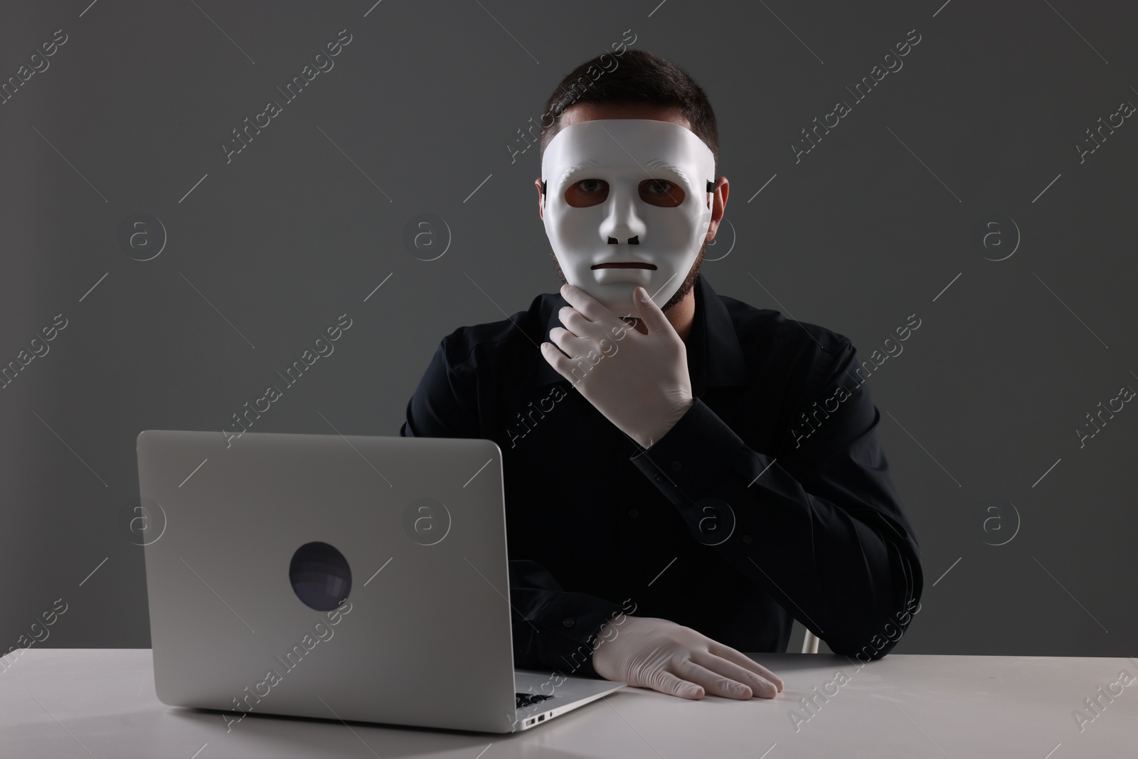 Photo of Man in mask and gloves sitting with laptop at white table against grey background