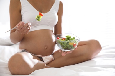 Photo of Young pregnant woman with bowl of vegetable salad in bedroom, closeup. Taking care of baby health