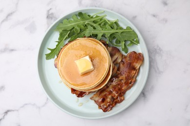 Photo of Tasty pancakes with butter, fried bacon and fresh arugula on white marble table, top view