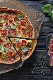 Cut delicious homemade quiche with prosciutto and ingredients on black wooden table, flat lay