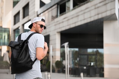 Handsome young man with stylish sunglasses and backpack near building outdoors, space for text