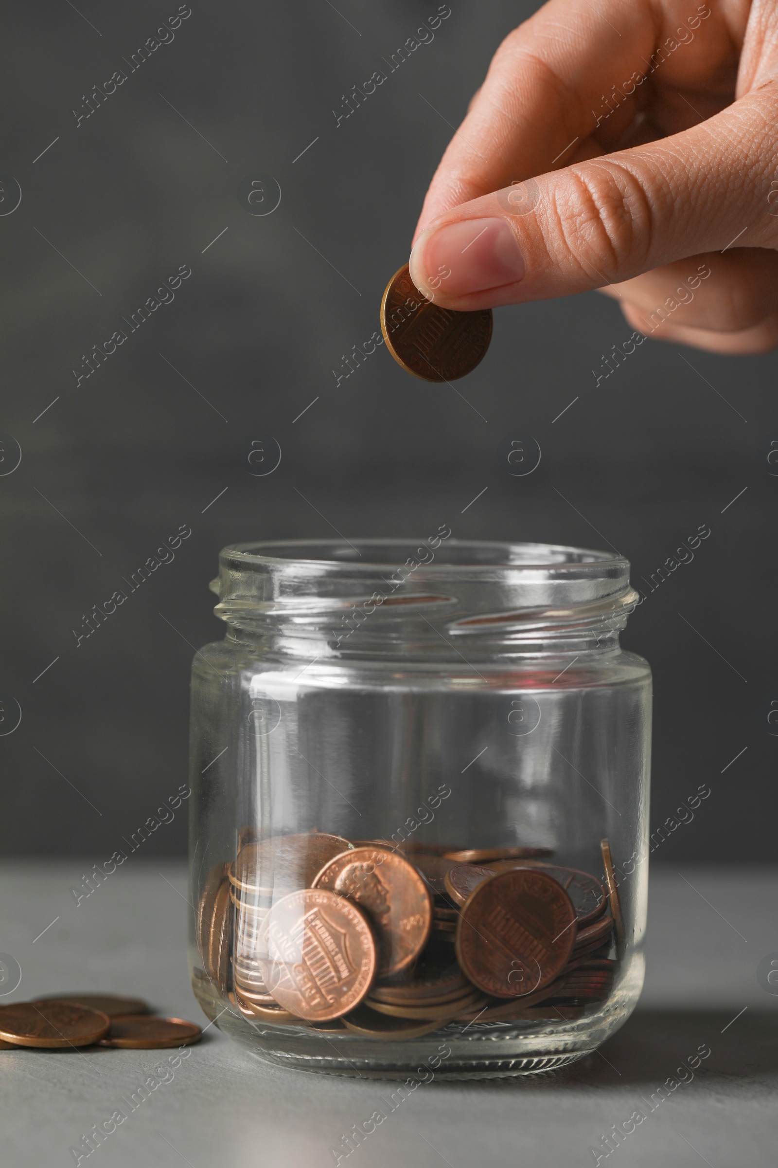 Photo of Woman putting coin in glass jar with money at grey table, closeup