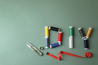 Photo of Abbreviation DIY of spools of colorful threads and sewing supplies on olive background, flat lay. Space for text