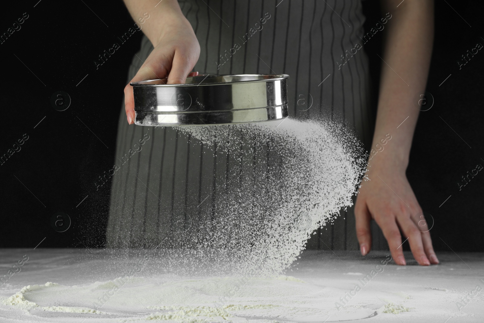 Photo of Woman sieving flour at table against black background, closeup