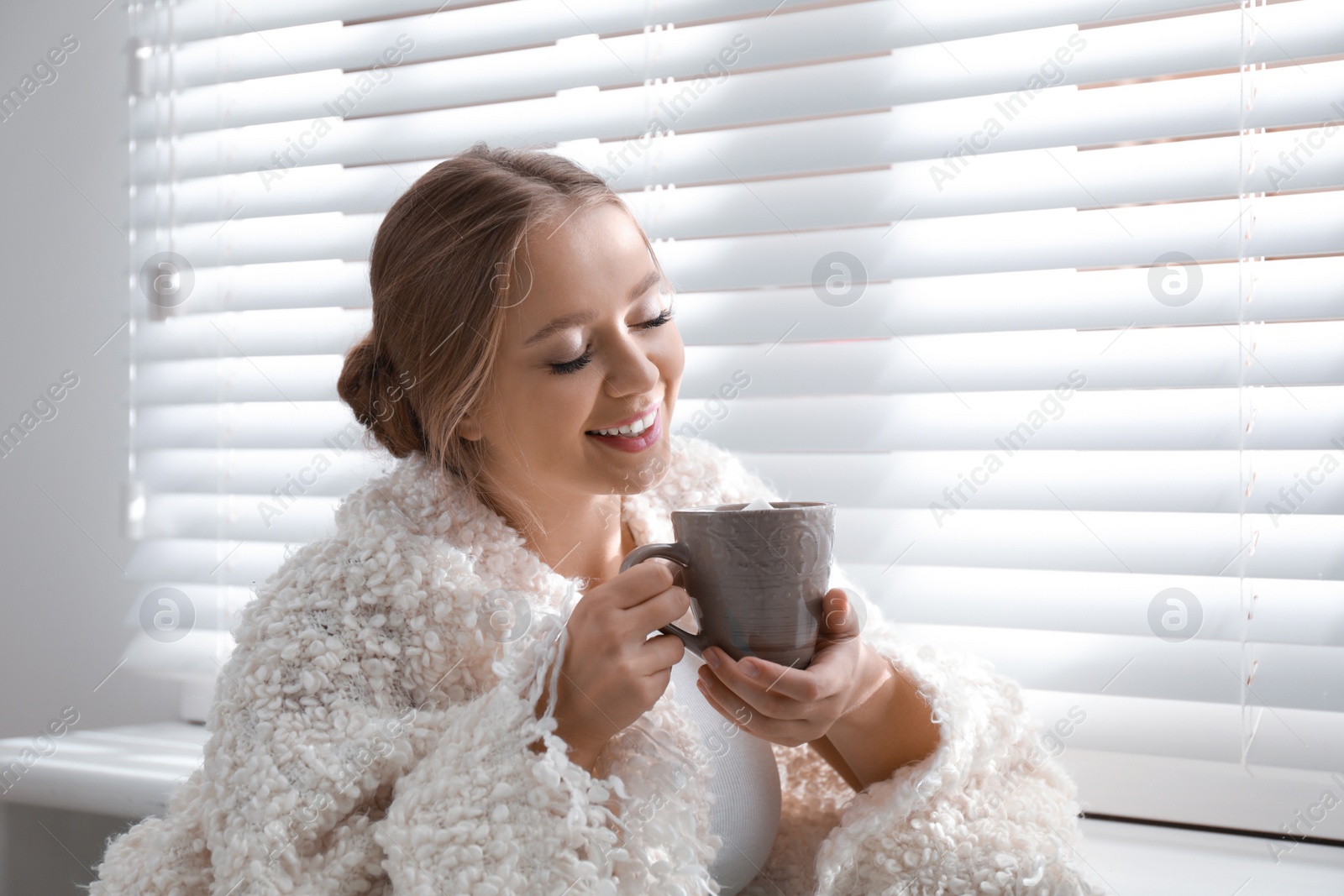 Photo of Beautiful young woman with cup of hot drink near window at home. Winter atmosphere