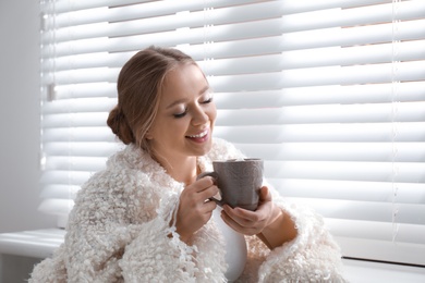 Beautiful young woman with cup of hot drink near window at home. Winter atmosphere