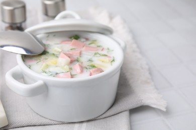 Delicious cold summer soup (okroshka) with boiled sausage in pot and spoon on white tiled table. Space for text