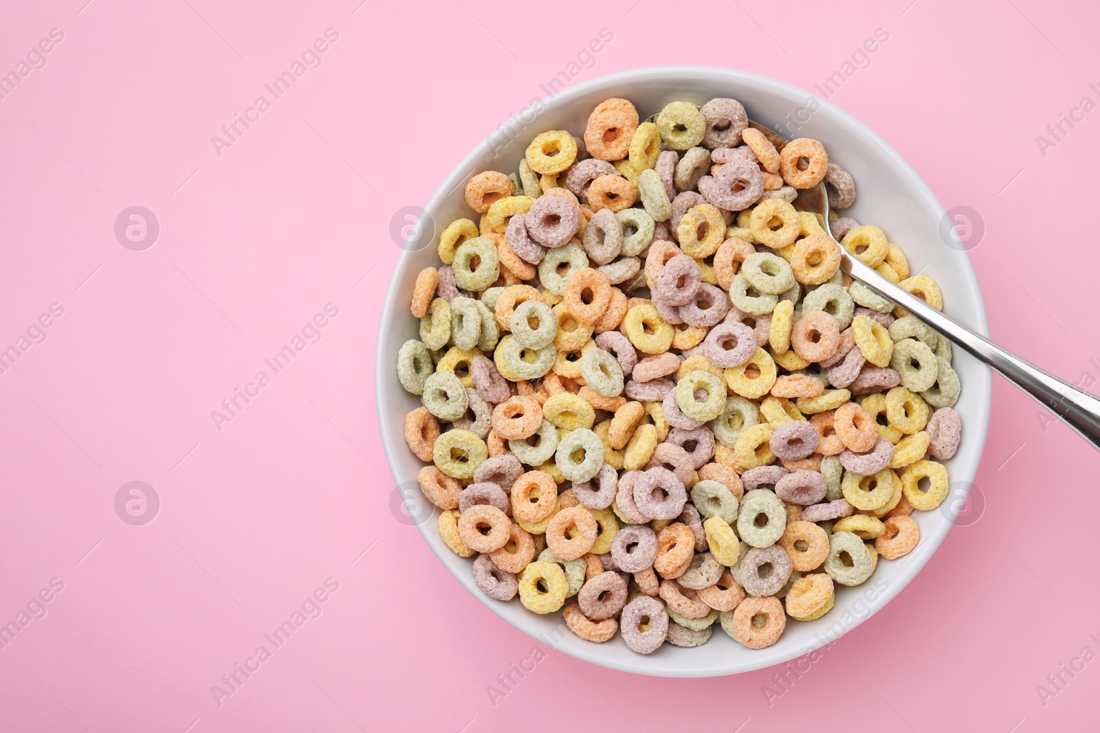 Photo of Tasty cereal rings in bowl and spoon on pink table, top view. Space for text