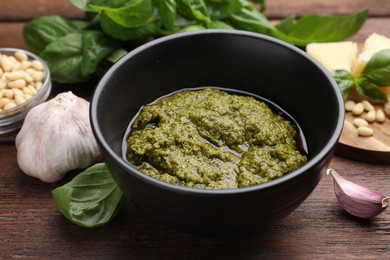 Photo of Tasty pesto sauce in bowl, basil, garlic and pine nuts on wooden table, closeup