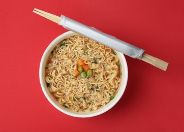 Cup of cooked instant noodles with chopsticks on color background, top view