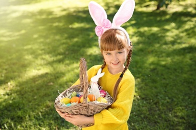 Photo of Cute little girl with bunny ears and basket of Easter eggs in park