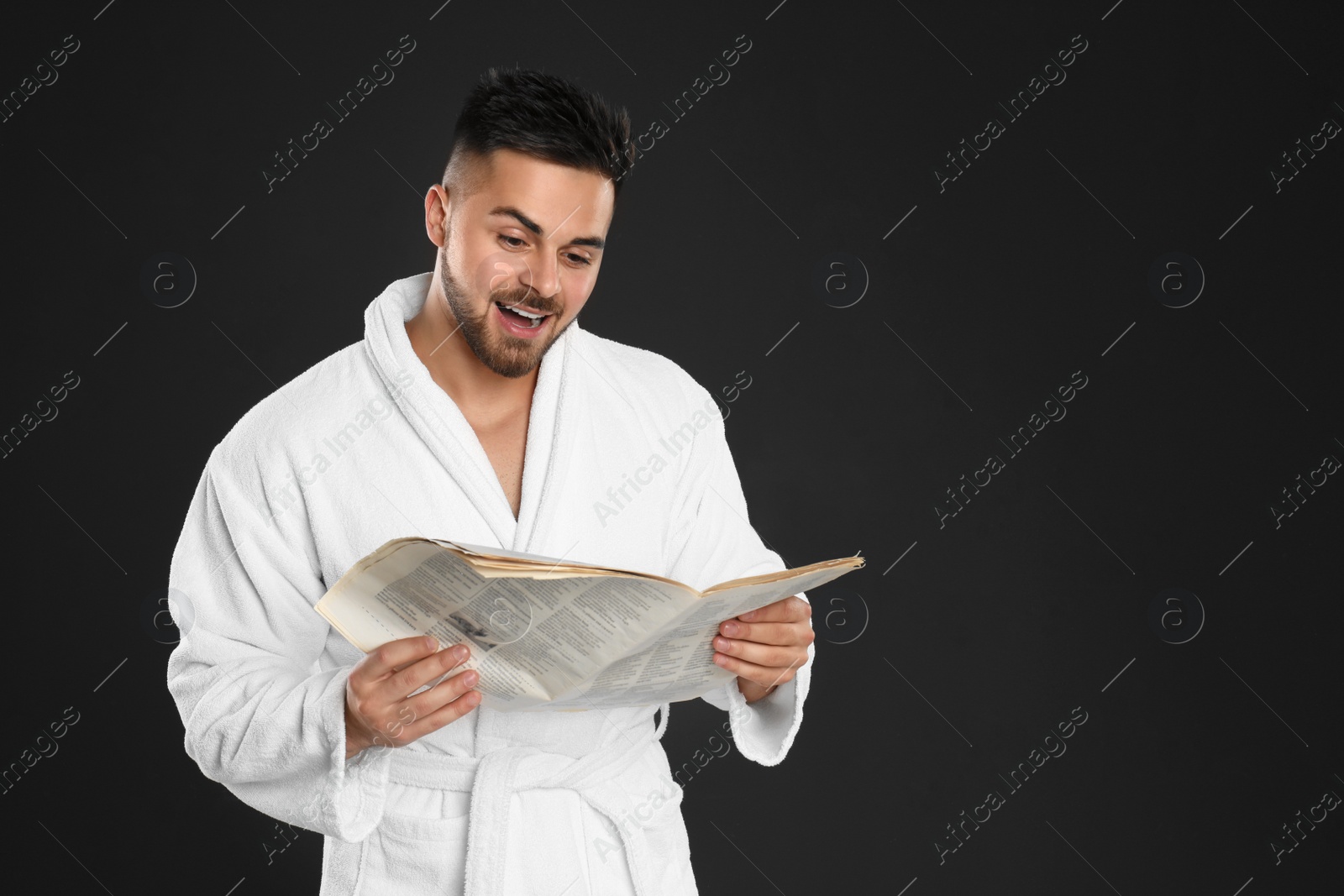 Photo of Young man in bathrobe reading newspaper on black background. Space for text