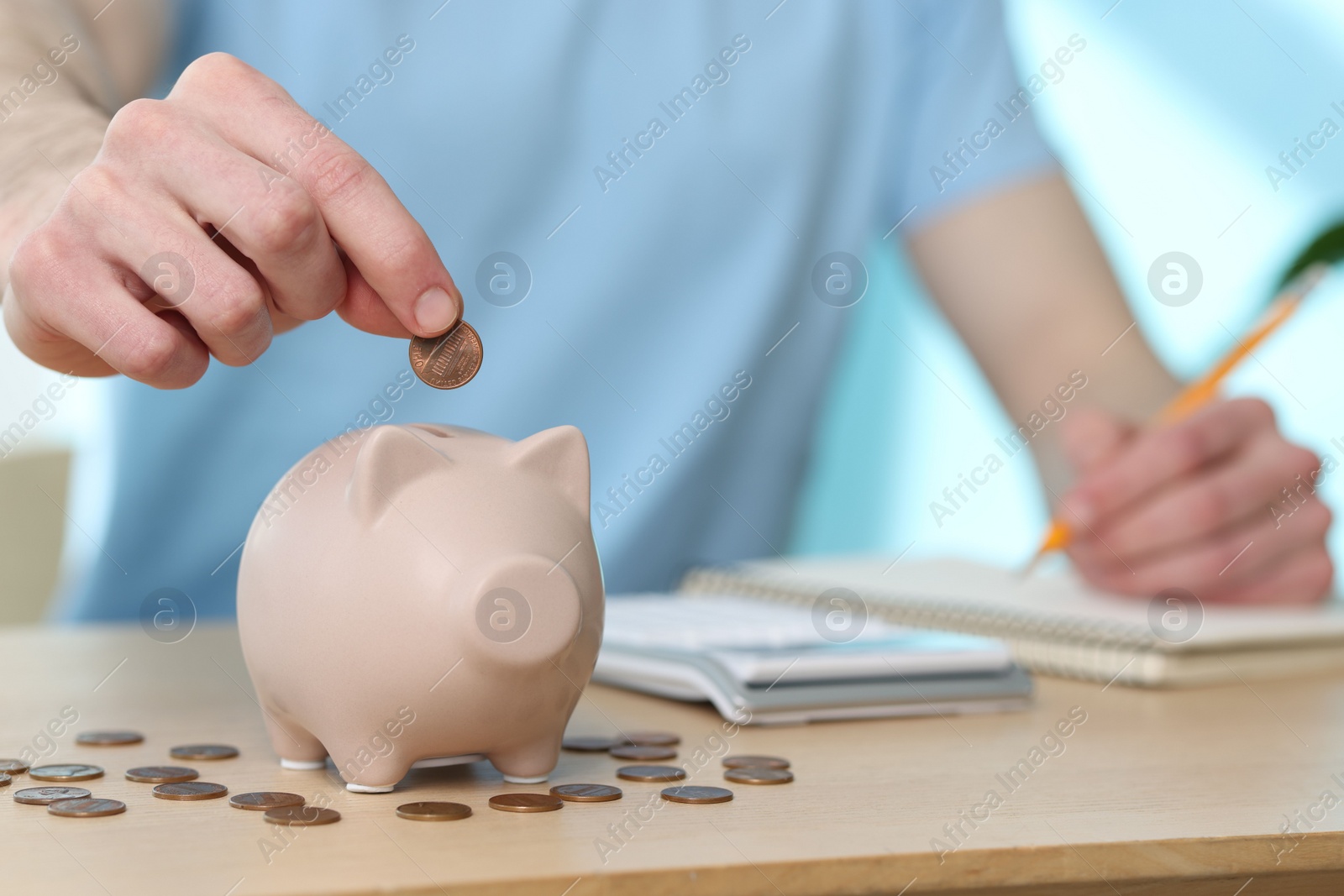 Photo of Financial savings. Man putting coin into piggy bank while writing down notes at wooden table, closeup